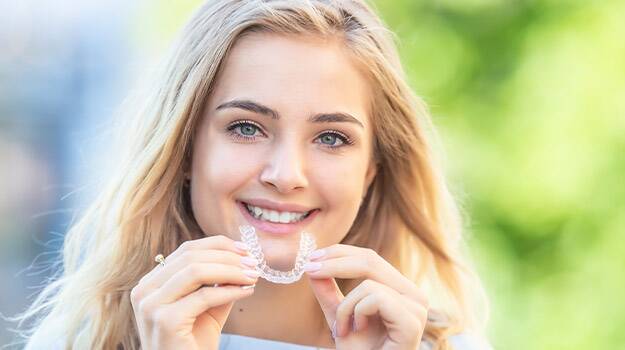A girl smiling while holding a clear aligner close to her mouth